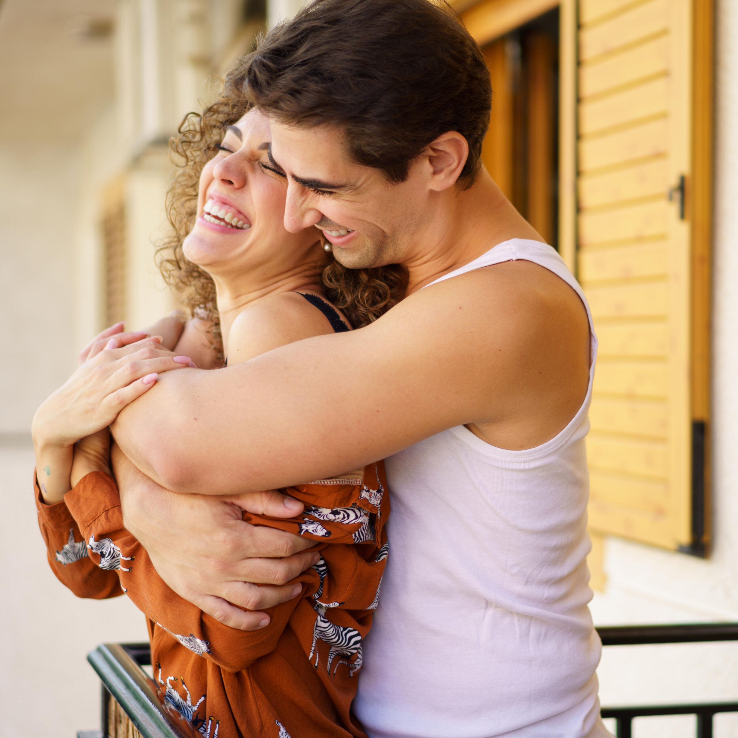 Happy couple hugging and laughing in daylight while standing at balcony